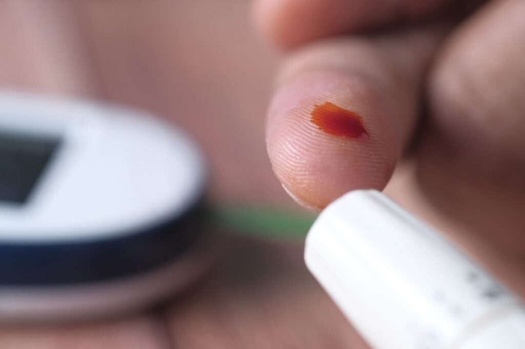 Ein Finger, mit einem kleinen Einstich, etwas Blut sowie ein Zuckermessstreifen